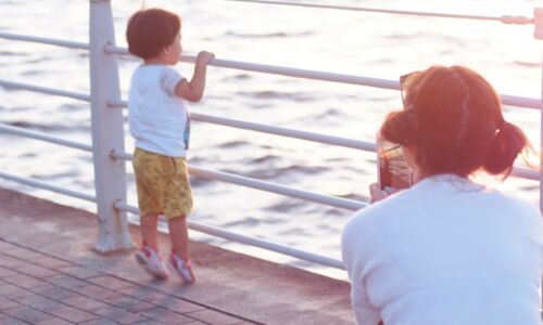 junger Mensch schaut aufs Meer und wird von älterem Menschen mit einem Smartphone fotografiert