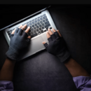 Draufsicht auf einen schwarzen Tisch: man sieht Hände mit schwarzen Fingerhandschuhen, die auf der Tastatur eines aufgeklappten Laptops liegen.