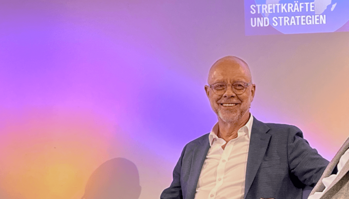 NDR Journalist Stefan Niemann sitzt lächelnd vor einer bunten Wandprojektion mit dem Titel "Streitkräfte und Strategien".
