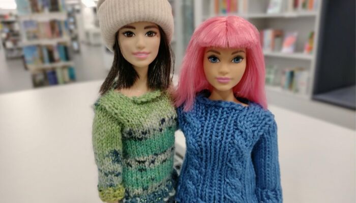 Zwei Puppen, eine mit Rosa Haaren und einem blauen Pullover und die andere mit braunen Haare, einer Mütze und einem grünen Pullover.