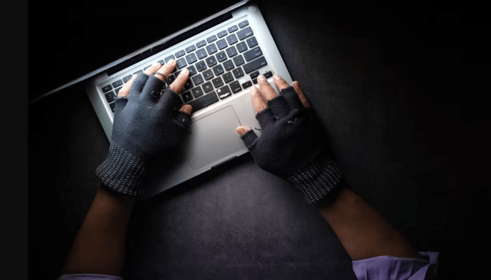 Draufsicht auf einen schwarzen Tisch: man sieht Hände mit schwarzen Fingerhandschuhen, die auf der Tastatur eines aufgeklappten Laptops liegen.