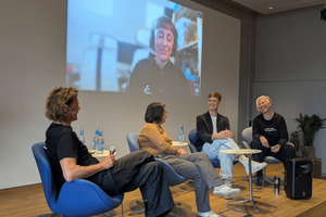 Vier Personen sitzen auf einem kleinen Podium und lächeln einander freundlich an. Hinter ihnen an der Wand eine Projektion einer Diskussionsteilnehmerin.