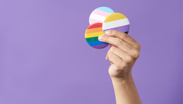 Eine Hand mit 3 Pride-Buttons in der Hand vor violettem Hintergrund.