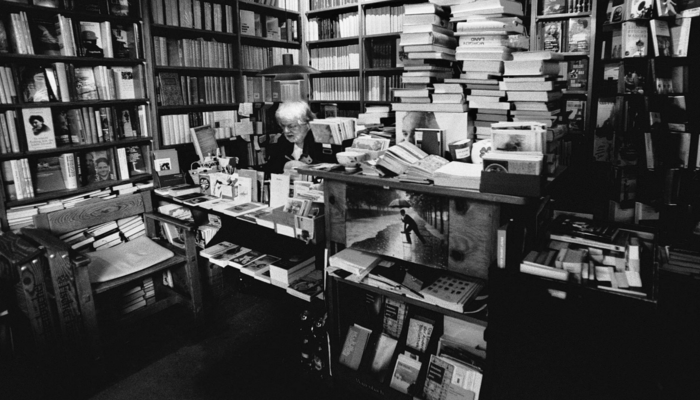 Schwarzweißfoto, auf dem ein älterer Mann zu sehen ist, der an der Kasse der Bücherstube Stolterfoht sitzt. Im Hintergrund stapeln sich viele Bücher, die Regale sind übervoll.