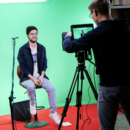 Ein junger Mann wird vor einem von einem Kameramann vor einem Greenscreen gefilmt. Er sitzt auf einem höhenverstellbaren Barhocker und seitlich rechts vor ihm ist ein kleines Richtmikrofon aufgebaut.