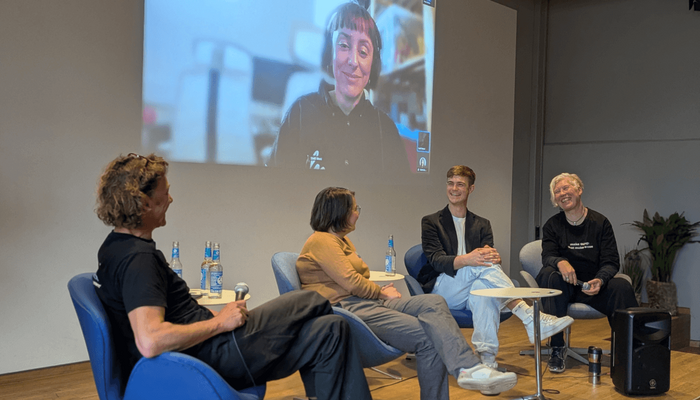Vier Personen sitzen auf einem kleinen Podium und lächeln einander freundlich an. Hinter ihnen an der Wand eine Projektion einer Diskussionsteilnehmerin.
