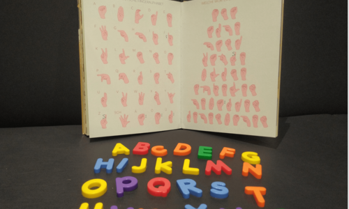 Ein Buch, dass das Fingeralphabet abbildet. Davor liegen bunte Buchstaben.