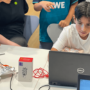 Kinder sitzen aufmerksam vor einem Laptop, an dem eine Leiterplatine von Makey Makey angeschlossen ist.
