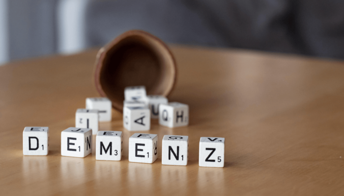Sechs nebeneinander aufgestellte Scrabble Spielwürfel liegen auf dem Tisch. Sie bilden das Wort "Demenz". Im Hintergrund unscharf: einige andere Würfel, die soeben aus einem umgefallenen Würfelbecher gefallen scheinen.