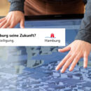 Weibliche Hände mit ausgestreckten Fingern über einem Screen, auf dem ein digitales Stadtplanungsmodell zu sehen ist.