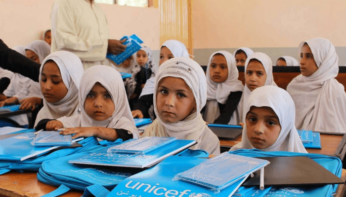 Mädchen mit weißem Hidschab sitzen nebeneinander in einem Klassenzimmer. Auf den Schultischen liegen hellblaue Unterrichtsmaterialien, die von UNICEF gespendet wurden. Im Hintergrund ist ein Lehrer mit weißem Hemd und weißer Gebetsmütze zu sehen.