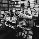 Schwarzweißfoto, auf dem ein älterer Mann zu sehen ist, der an der Kasse der Bücherstube Stolterfoht sitzt. Im Hintergrund stapeln sich viele Bücher, die Regale sind übervoll.