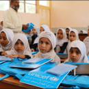 Mädchen mit weißem Hidschab sitzen nebeneinander in einem Klassenzimmer. Auf den Schultischen liegen hellblaue Unterrichtsmaterialien, die von UNICEF gespendet wurden. Im Hintergrund ist ein Lehrer mit weißem Hemd und weißer Gebetsmütze zu sehen.