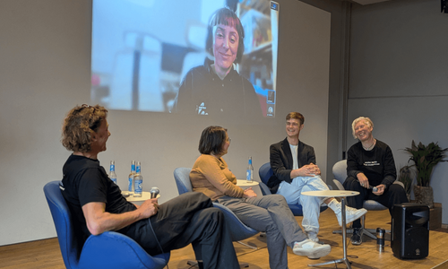 Vier Personen sitzen auf einem kleinen Podium und lächeln einander freundlich an. Hinter ihnen an der Wand eine Projektion einer Diskussionsteilnehmerin.