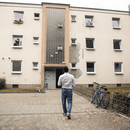 Rückensansicht eines Mannes, der sich dem Eingang eines Mehrfamilienhauses mit heruntergekommener Fassade nähert.