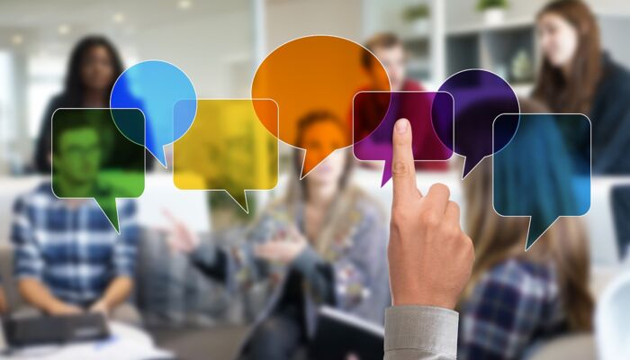 Im Vordergrund ist ein gestreckter Zeigefinger zu sehen. Im Hintergrund etwas unscharf mehrere Diskussionsteilnehmer*innen. Verschiedene bunte, transparente Sprechblasen liegen teilweise über den Gesichtern.