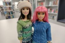 Zwei Puppen, eine mit Rosa Haaren und einem blauen Pullover und die andere mit braunen Haare, einer Mütze und einem grünen Pullover.
