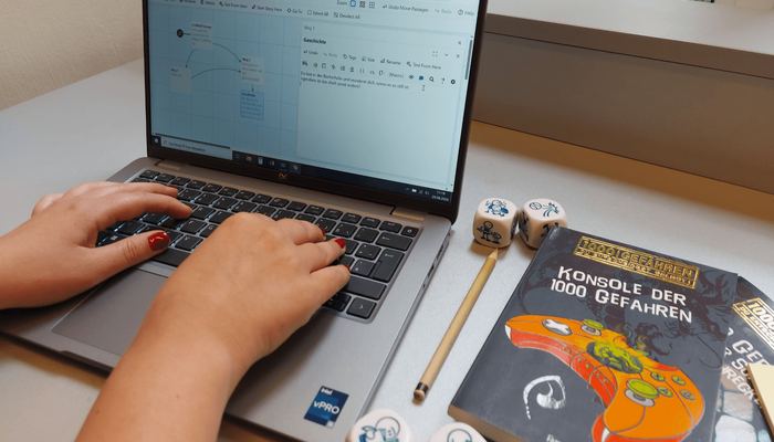An einem Laptop wird ein Computerspiel entworfen. Rechts neben dem Laptop liegt ein Buch und Würfel.