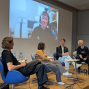 Vier Personen sitzen auf einem kleinen Podium und lächeln einander freundlich an. Hinter ihnen an der Wand eine Projektion einer Diskussionsteilnehmerin.