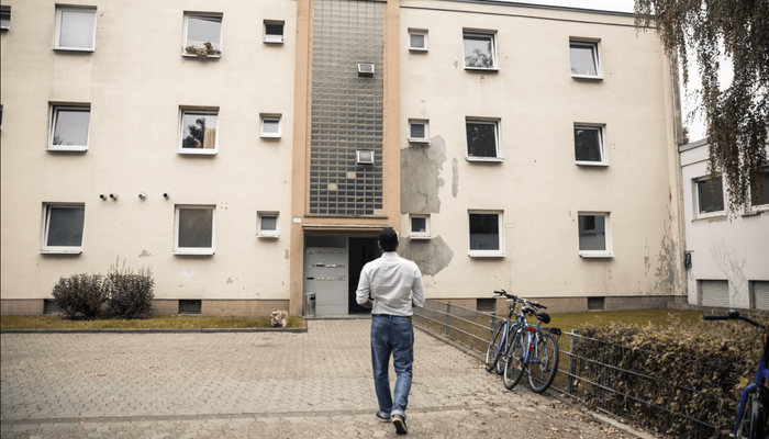 Rückensansicht eines Mannes, der sich dem Eingang eines Mehrfamilienhauses mit heruntergekommener Fassade nähert.