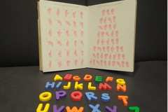 Ein Buch, dass das Fingeralphabet abbildet. Davor liegen bunte Buchstaben.