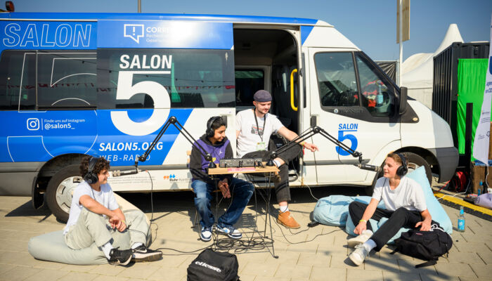 Foto zeigt 4 junge Personen vor einem Betriebsbus, die eine offenbar eine Audiosendung aufzeichnen