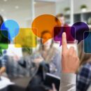 Im Vordergrund ist ein gestreckter Zeigefinger zu sehen. Im Hintergrund etwas unscharf mehrere Diskussionsteilnehmer*innen. Verschiedene bunte, transparente Sprechblasen liegen teilweise über den Gesichtern.