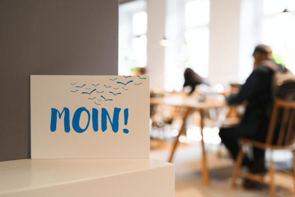 Postkarte mit der Aufschrift „Moin!“ vor Menschen an Leseplätzen in einer Bücherhalle