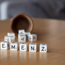 Sechs nebeneinander aufgestellte Scrabble Spielwürfel liegen auf dem Tisch. Sie bilden das Wort "Demenz". Im Hintergrund unscharf: einige andere Würfel, die soeben aus einem umgefallenen Würfelbecher gefallen scheinen.