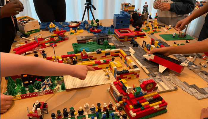 Kinder stehen um einen Tisch herum, auf dem ein Flughafen aus LEGO aufgebaut wurde. Im Hintergrund ist ein IPad ist auf einem Stativ als Kamera auf den Tisch gerichtet.