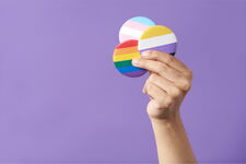 Eine Hand mit 3 Pride-Buttons in der Hand vor violettem Hintergrund.