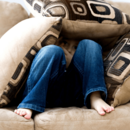 Foto von einem kleinen Kind, welches sich auf einer beigefarbenen Couch unter mehreren Kissen versteckt hat. Es sind lediglich die Beine des Kindes und im Hintergrund Augen und Hände des Kindes zu sehen.