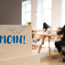 Postkarte mit der Aufschrift „Moin!“ vor Menschen an Leseplätzen in einer Bücherhalle