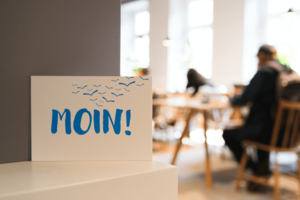 Postkarte mit der Aufschrift „Moin!“ vor Menschen an Leseplätzen in einer Bücherhalle