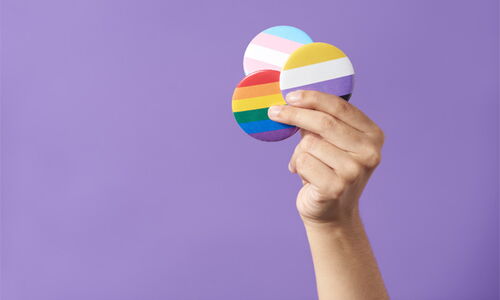 Eine Hand mit 3 Pride-Buttons in der Hand vor violettem Hintergrund.