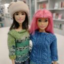 Zwei Puppen, eine mit Rosa Haaren und einem blauen Pullover und die andere mit braunen Haare, einer Mütze und einem grünen Pullover.