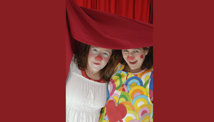 Zwei Frauen, die als Clowns geschminkt sind, schauen unter einem roten Vorhang heraus.