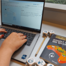 An einem Laptop wird ein Computerspiel entworfen. Rechts neben dem Laptop liegt ein Buch und Würfel.