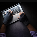 Draufsicht auf einen Laptop, auf dem eine Person mit fingerfreien, schwarzen Handschuhen tippt. Die Umgebung ist dunkel, nur der Bildschirm des Laptops spendet Licht.