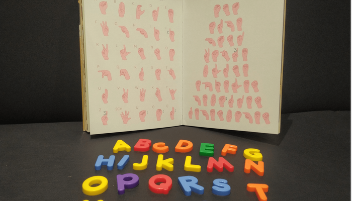 Ein Buch, dass das Fingeralphabet abbildet. Davor liegen bunte Buchstaben.