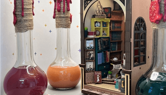 Vor einem Miniatur-Bookstore stehen 3 große Kolbengefäße. die mit unterschiedlichen Flüssigkeiten gefüllt sind. Im Vordergrund liegen einzelne Spielkarten.