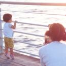 junger Mensch schaut aufs Meer und wird von älterem Menschen mit einem Smartphone fotografiert
