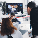 Eine Person blickt einer anderen an einem Scheibtisch mit Laptops über die Schulter