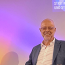 NDR Journalist Stefan Niemann sitzt lächelnd vor einer bunten Wandprojektion mit dem Titel "Streitkräfte und Strategien".