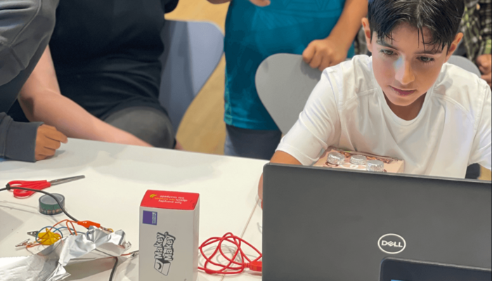 Kinder sitzen aufmerksam vor einem Laptop, an dem eine Leiterplatine von Makey Makey angeschlossen ist.
