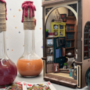 Vor einem Miniatur-Bookstore stehen 3 große Kolbengefäße. die mit unterschiedlichen Flüssigkeiten gefüllt sind. Im Vordergrund liegen einzelne Spielkarten.