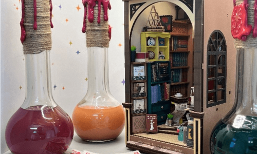 Vor einem Miniatur-Bookstore stehen 3 große Kolbengefäße. die mit unterschiedlichen Flüssigkeiten gefüllt sind. Im Vordergrund liegen einzelne Spielkarten.