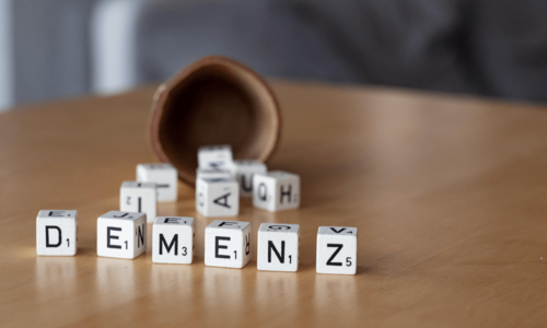 Sechs nebeneinander aufgestellte Scrabble Spielwürfel liegen auf dem Tisch. Sie bilden das Wort "Demenz". Im Hintergrund unscharf: einige andere Würfel, die soeben aus einem umgefallenen Würfelbecher gefallen scheinen.