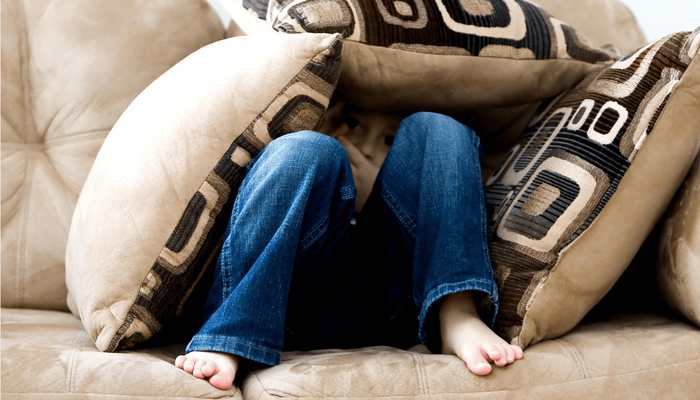 Foto von einem kleinen Kind, welches sich auf einer beigefarbenen Couch unter mehreren Kissen versteckt hat. Es sind lediglich die Beine des Kindes und im Hintergrund Augen und Hände des Kindes zu sehen.