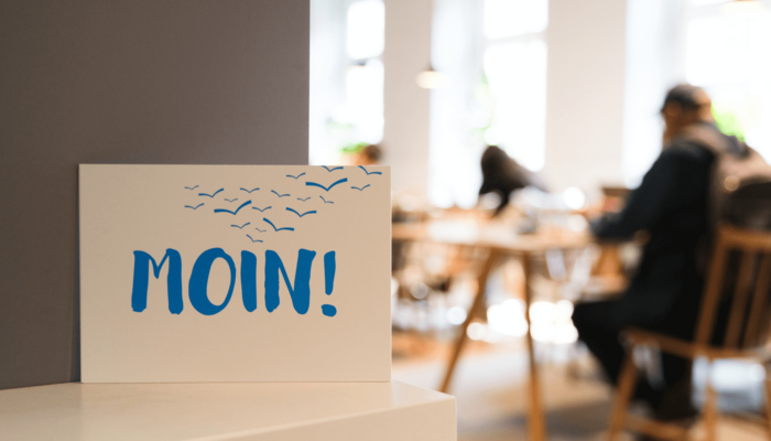 Postkarte mit der Aufschrift „Moin!“ vor Menschen an Leseplätzen in einer Bücherhalle
